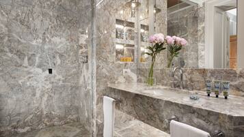 A bathroom with a marble sink, a mirror, a shower, and a vase with flowers.
