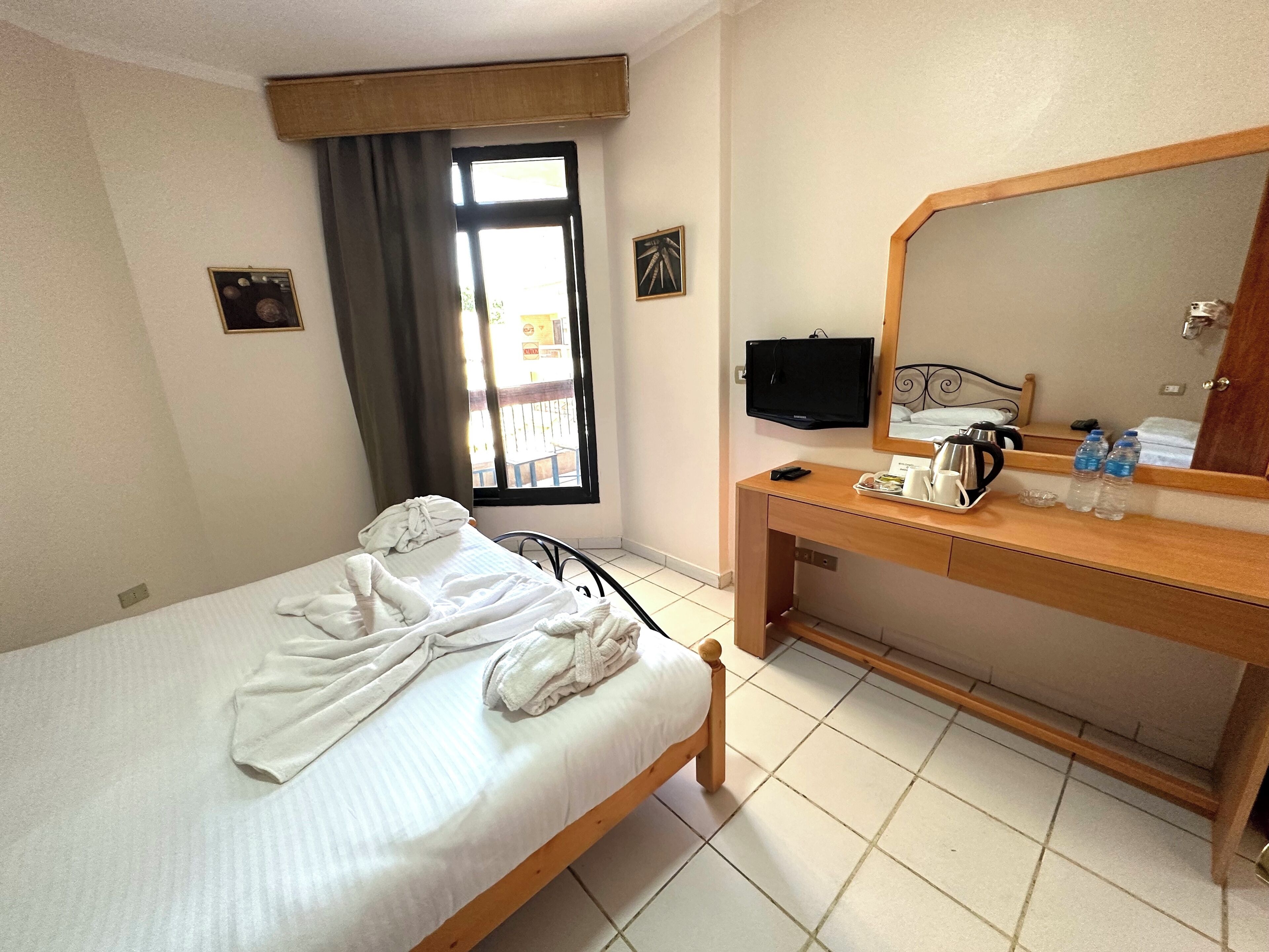 A hotel room with a bed, a wooden desk, a flat-screen TV, a mirror, and a window with curtains.