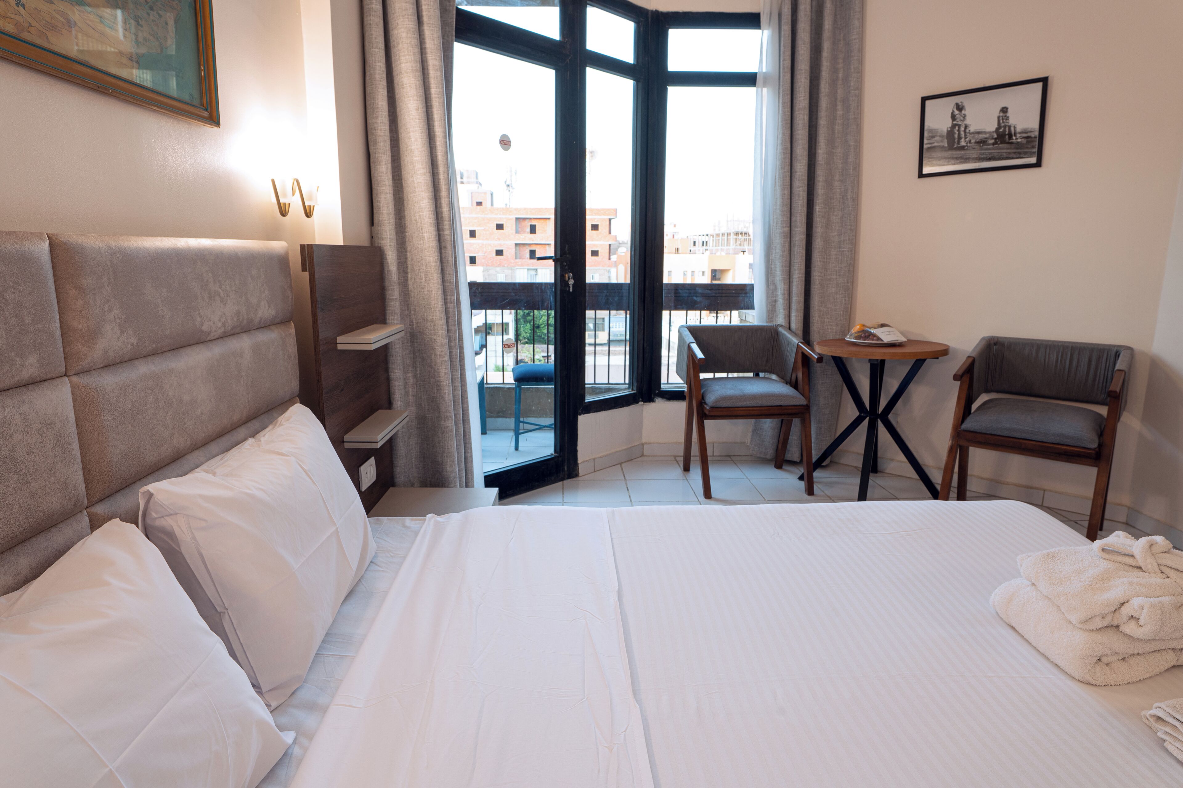 A hotel room with a large bed, two chairs, a small table, and a balcony with a view of buildings.