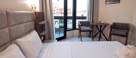 A hotel room with a large bed, two chairs, a small table, and a balcony with a view of buildings.