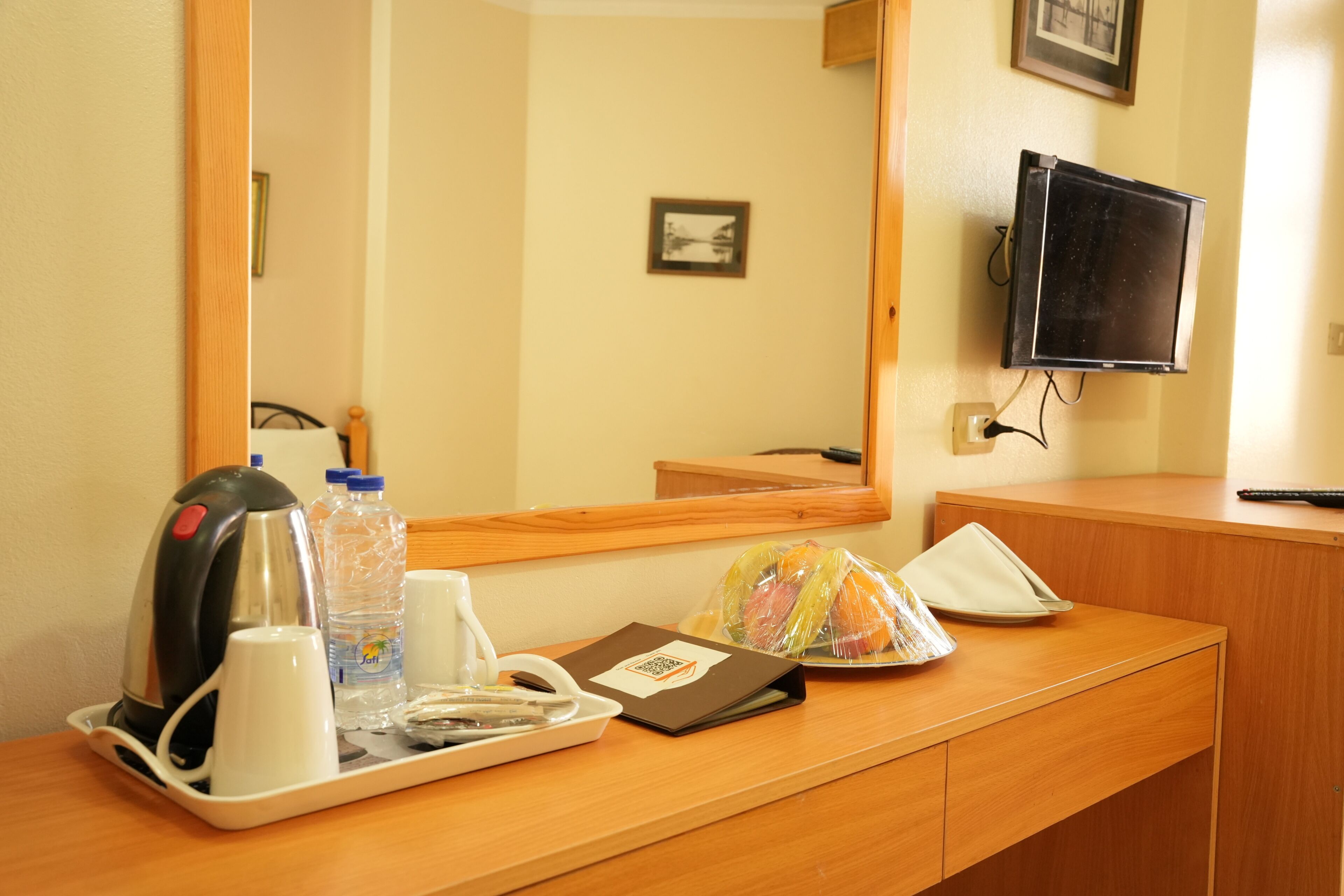 A hotel room with a wooden desk, a kettle, a water bottle, a tray with a wrapped fruit, a remote control, and a television mounted on the wall.