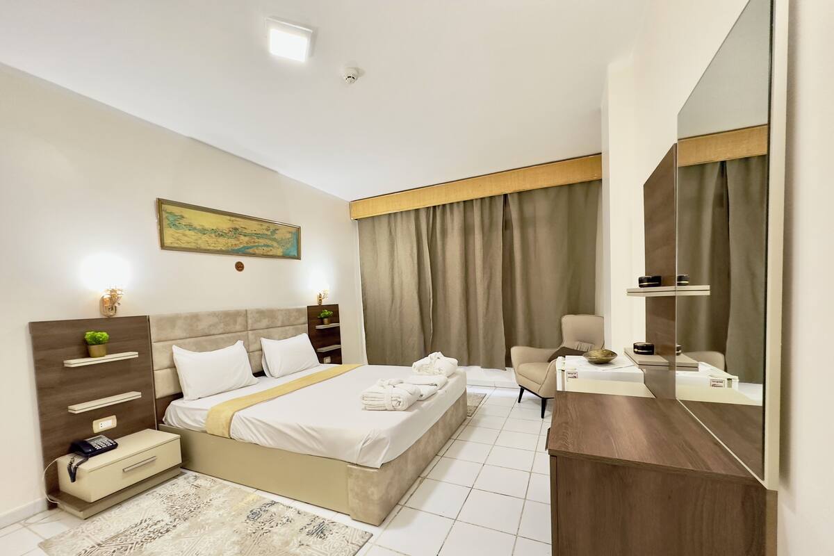 A modern hotel room with a bed, bedside tables, a desk, a chair, a large mirror, and a painting on the wall.