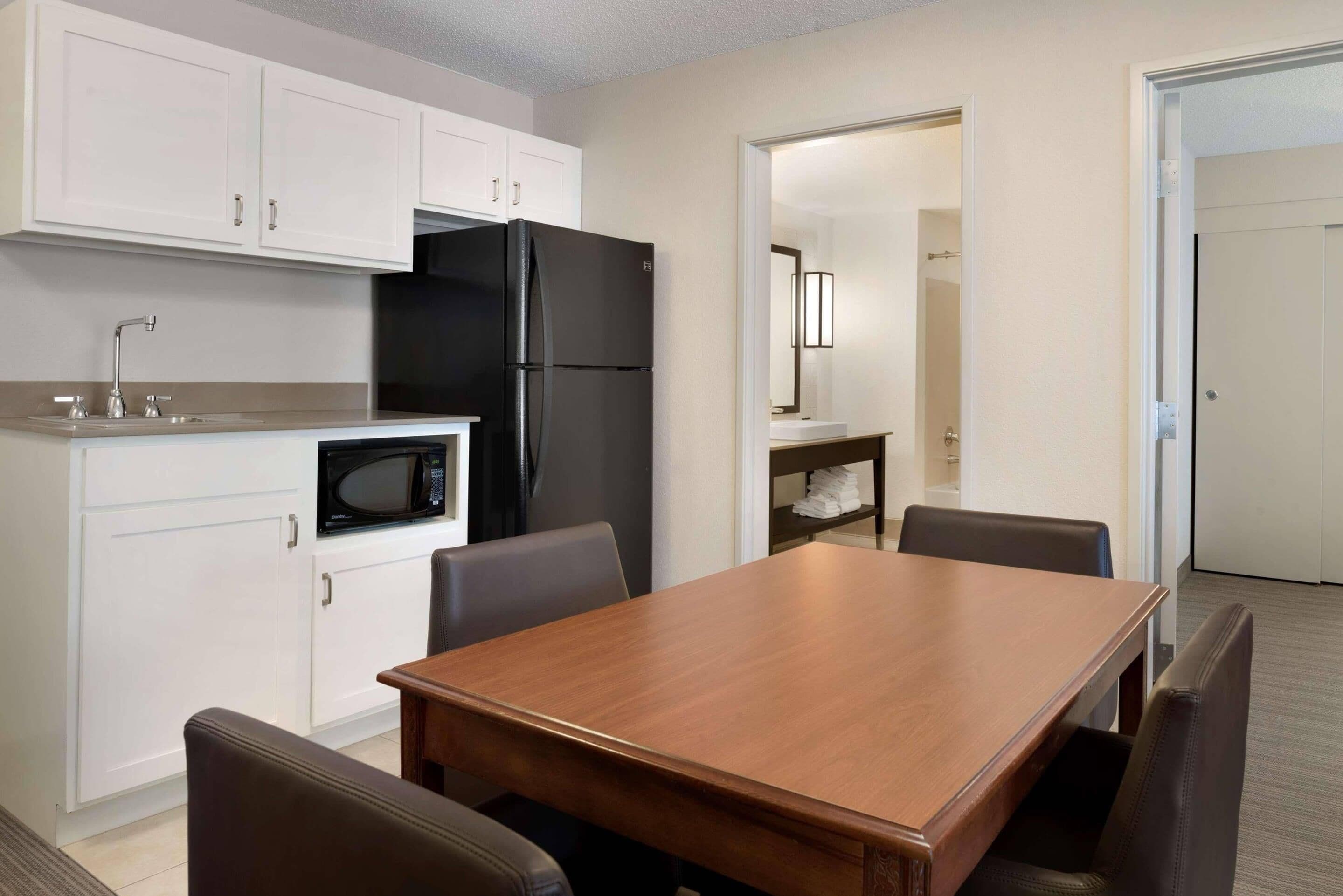 Cocina compacta con armarios blancos, un refrigerador negro, un microondas y una mesa de comedor de madera con cuatro sillas.