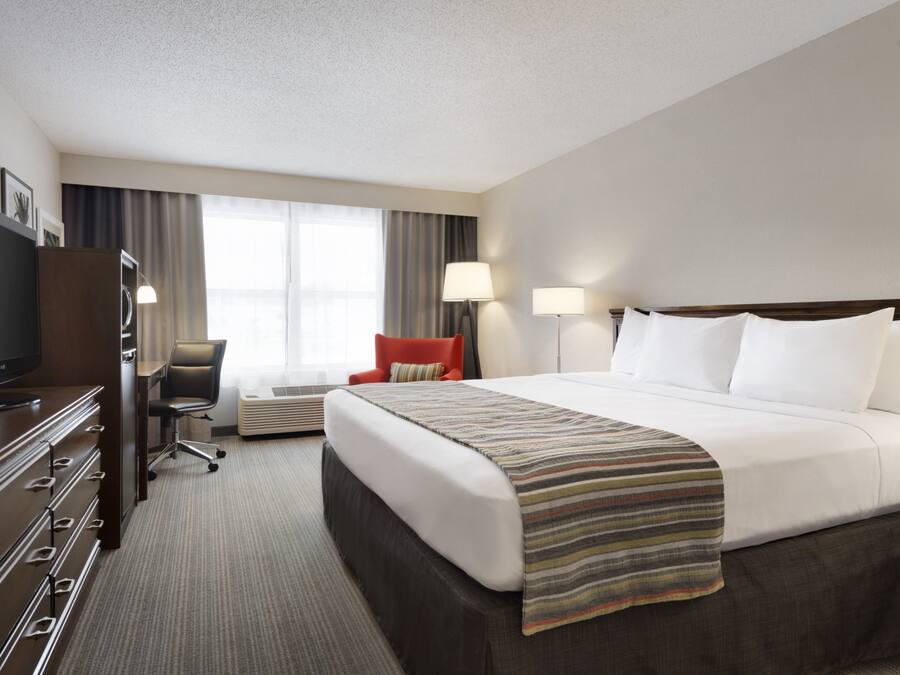 A hotel room with a large bed, a desk with a flat-screen TV, a chair, two bedside lamps, and a window with curtains.