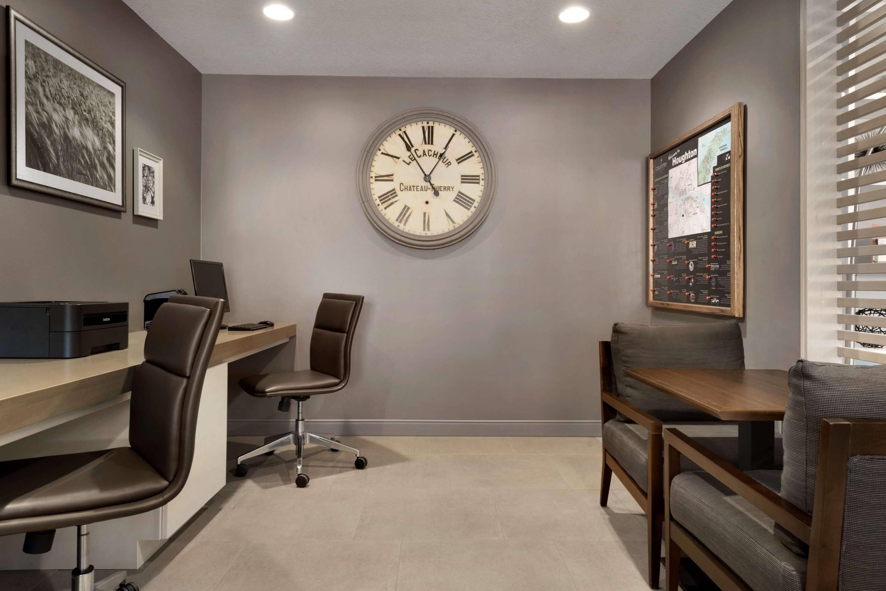 A modern office space with a large wall clock, a desk with a computer, a chair, and a small table with a chair.