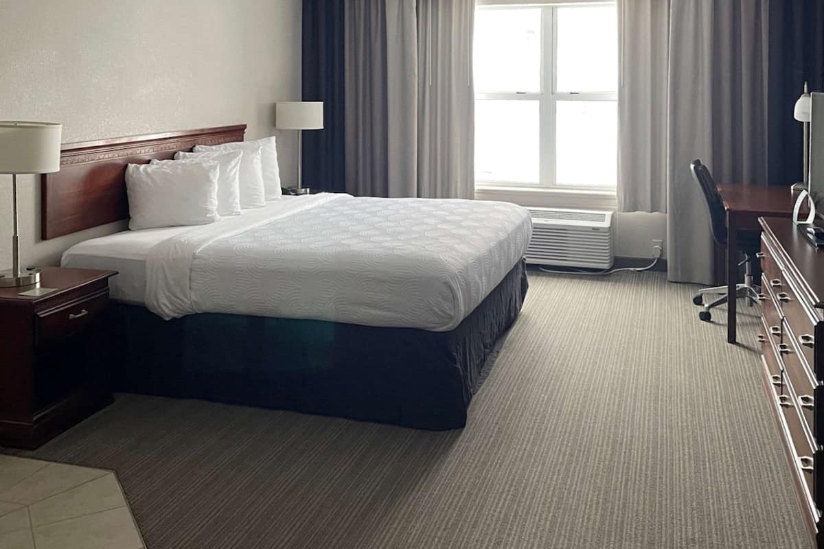 Una habitación de hotel con una cama grande, un escritorio, una silla, una lámpara y un ventanal con cortinas.