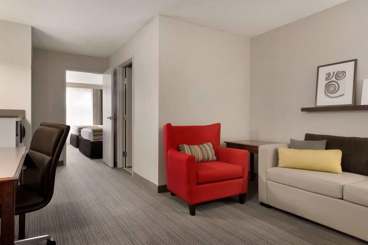 Habitación de hotel con un sillón rojo, un sofá color beige, una mesita y un escritorio con silla. Se ve una puerta que da a un pasillo con cama y ventana.