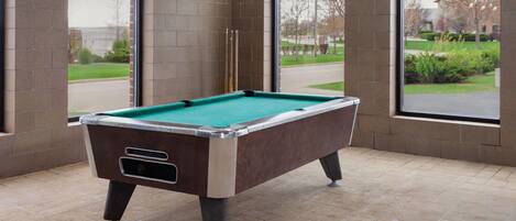 A pool table in a room with large windows, brick walls, and a tiled floor.