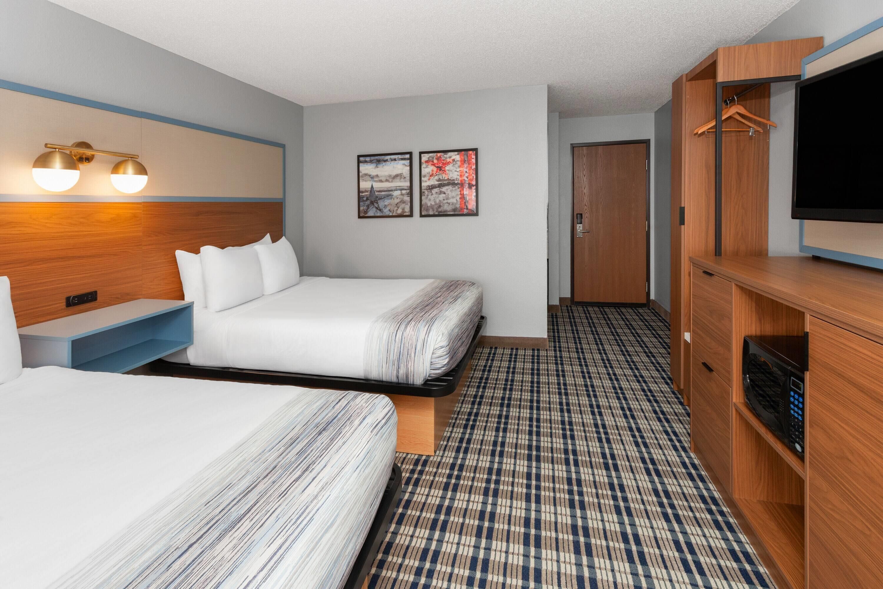 A hotel room with two beds, a flat-screen TV, a wooden wardrobe, and a wall-mounted light fixture.