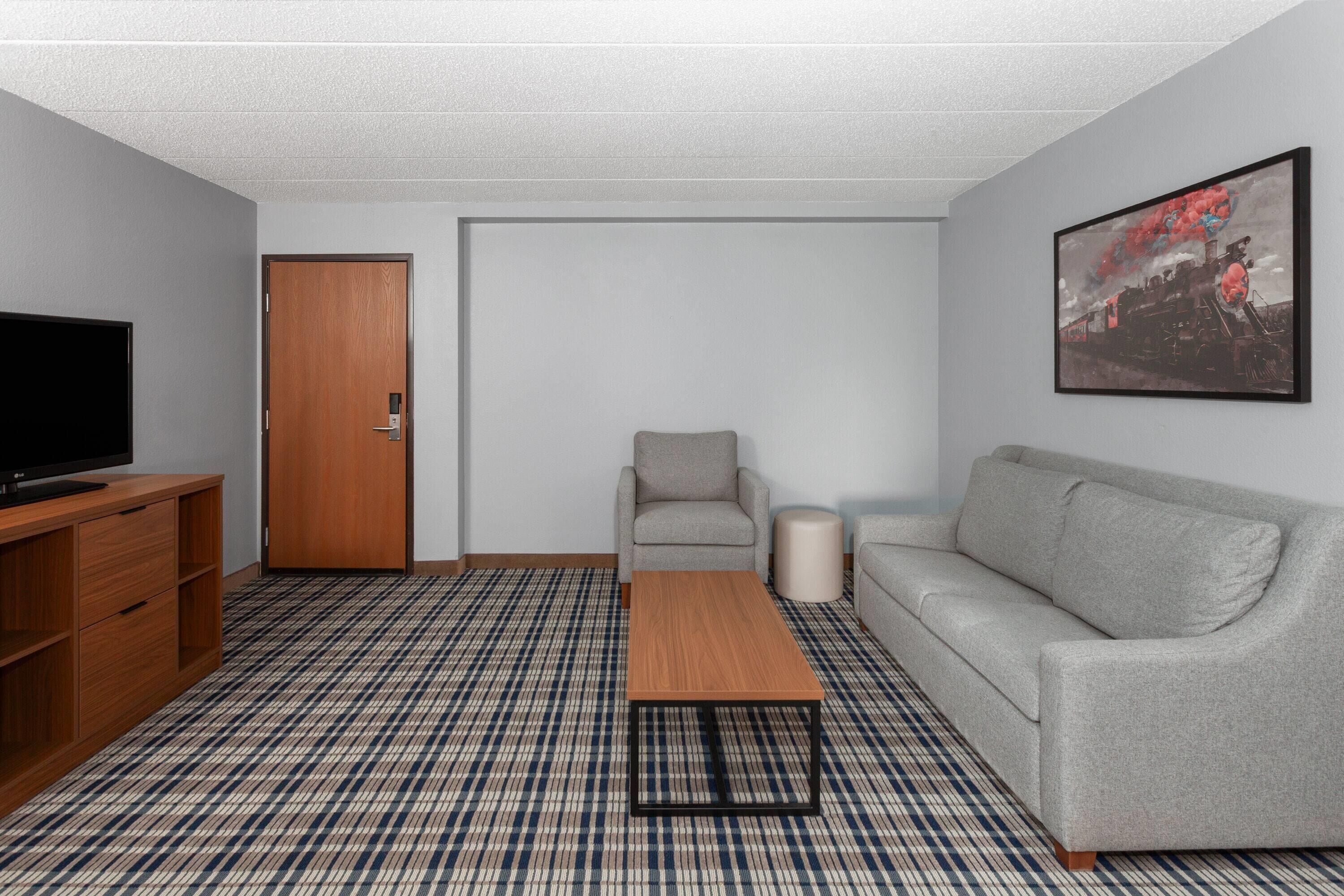 A room with a television, a wooden cabinet, a plaid carpet, a grey sofa, a small armchair, and a painting on the wall.