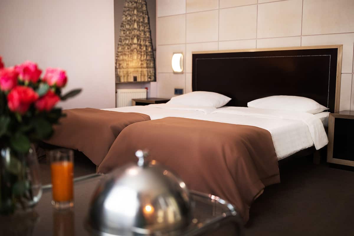 A hotel room with two beds, a bedside table with a glass of orange juice, and a vase with pink flowers.