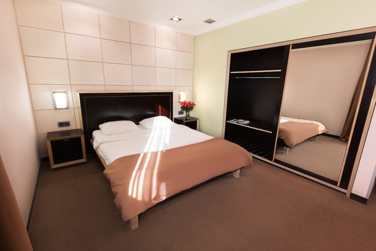 A hotel room with a large bed, a bedside table with a lamp, a nightstand, and a wardrobe with a mirror.