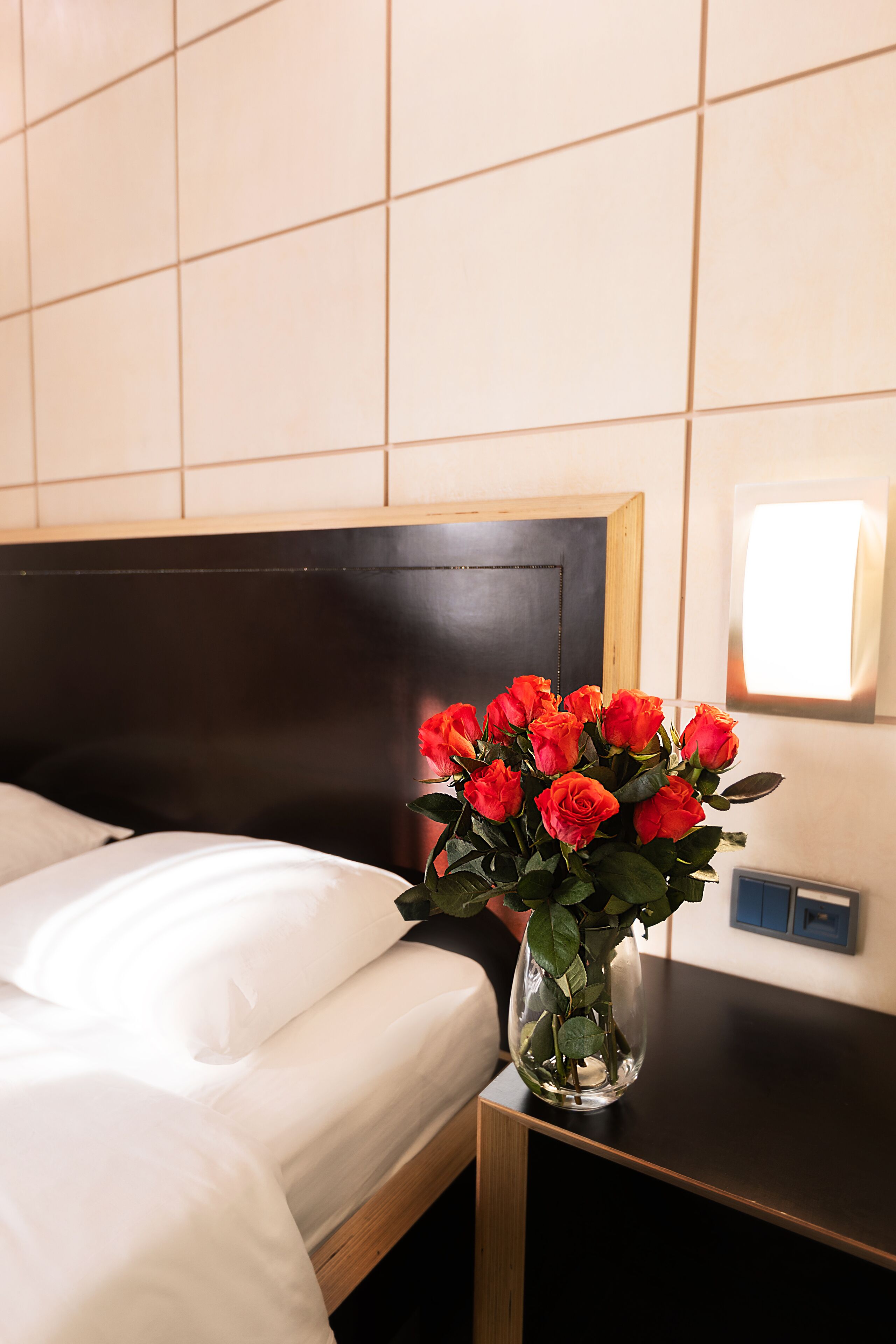 A hotel room with a bed, a nightstand, and a vase with red flowers.