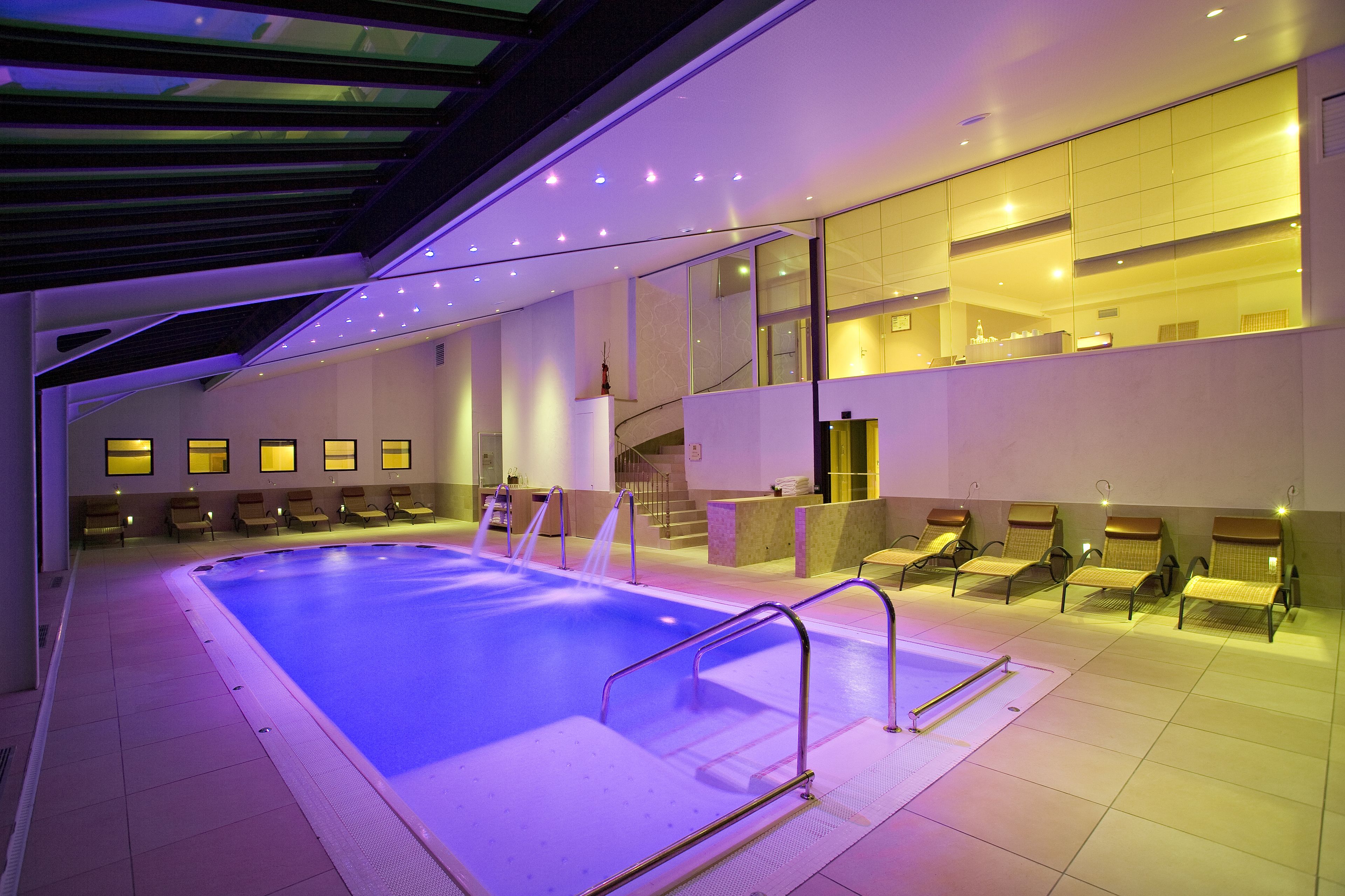 Una piscina interior moderna con techo de vidrio, varias sillas de descanso y una escalera que conduce a niveles superiores.