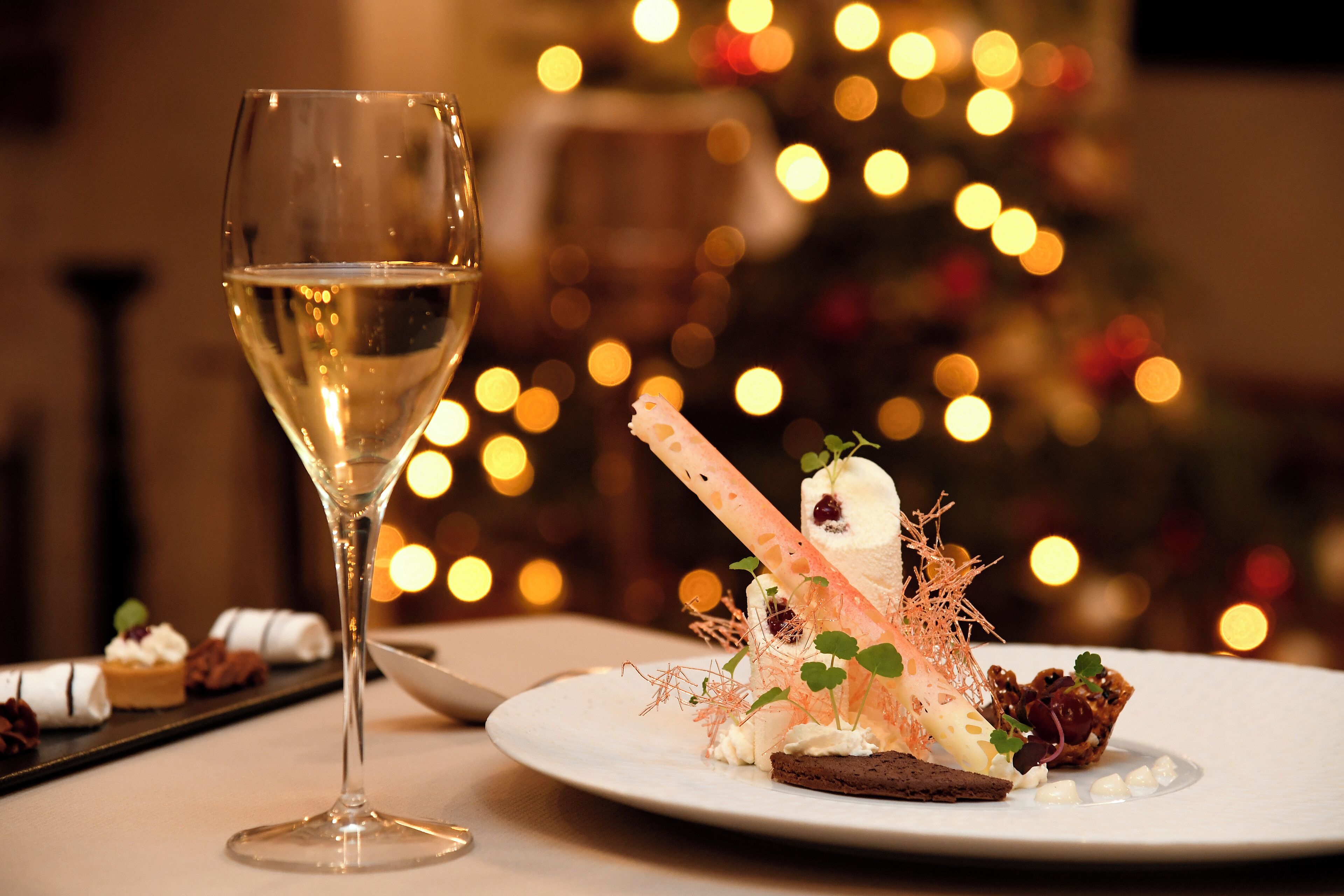 Una copa de vino blanco, un plato de postres con varios dulces y un fondo desenfocado con luces.
