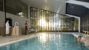 A modern indoor swimming pool with clear glass walls, a sunlit pool area, and a lounge area with sunbeds.
