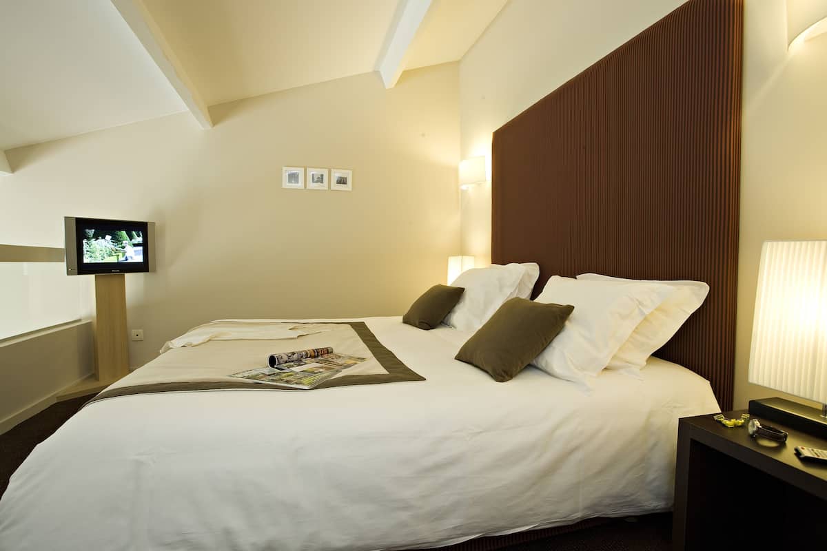 Habitación de hotel con una cama grande, un televisor de pantalla plana montado en la pared, una mesita de noche con lámpara y una ventana con persianas.