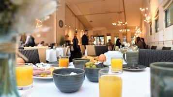 Un desayuno con una mesa dispuesta para dos personas, con platos de comida, tazones, vasos de zumo de naranja y un jarrón con flores.