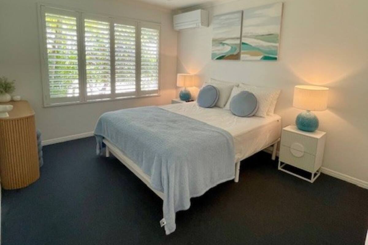 A bedroom with a bed, bedside lamps, a ceiling fan, a window with blinds, and a wall-mounted painting.