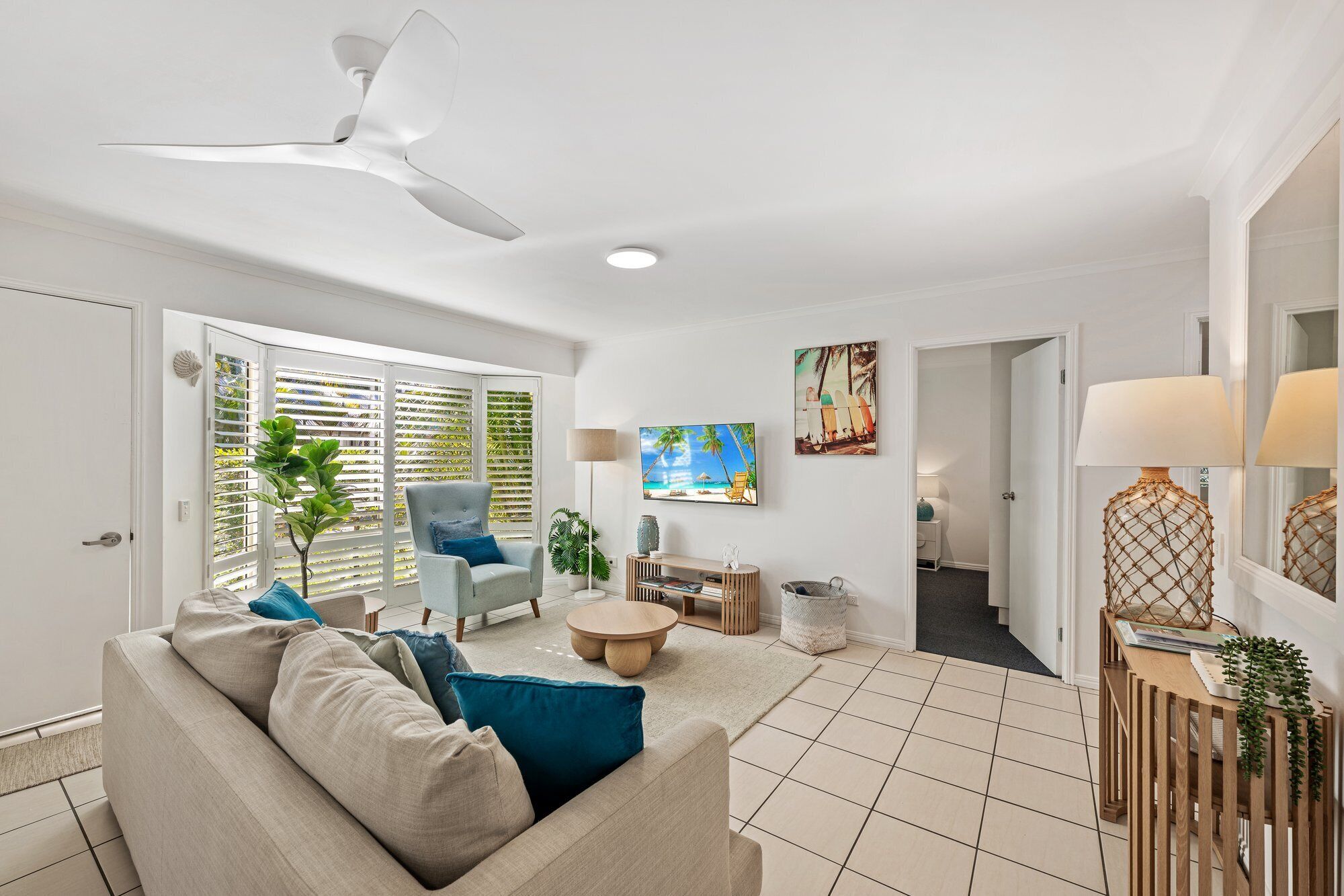 A modern living room with a ceiling fan, a sofa with cushions, a coffee table, a TV, and a painting of a beach scene.