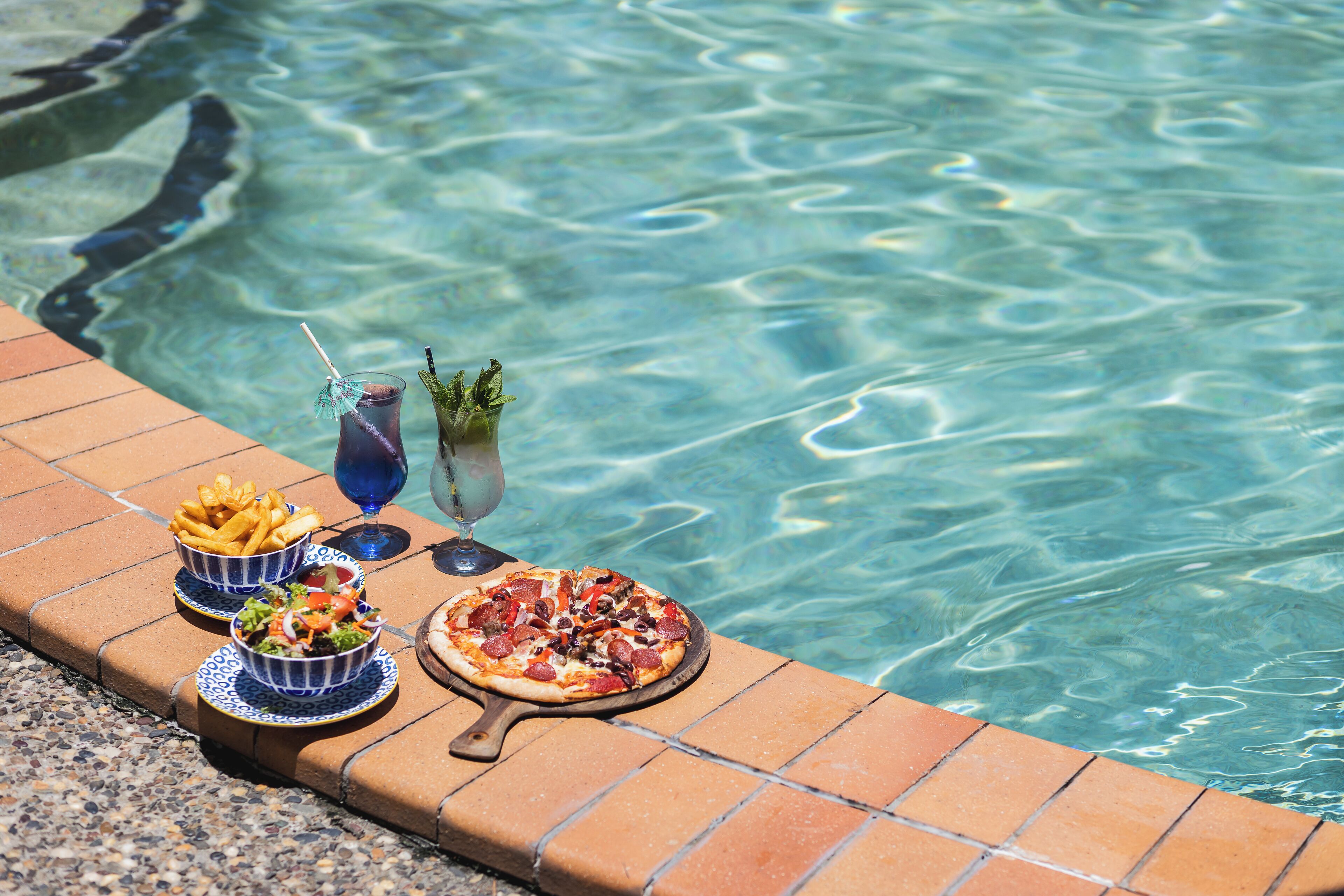 Mesa junto a la piscina con pizza, ensalada y bebidas.