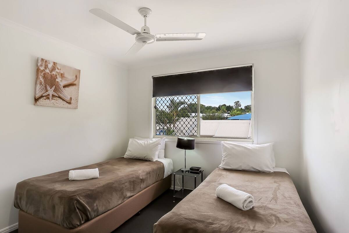 A room with two beds, a ceiling fan, a window with a view, and a painting on the wall.