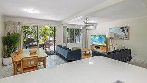 Una sala de estar moderna con comedor, un sofá, un televisor y un amplio ventanal con vistas a la vegetación.