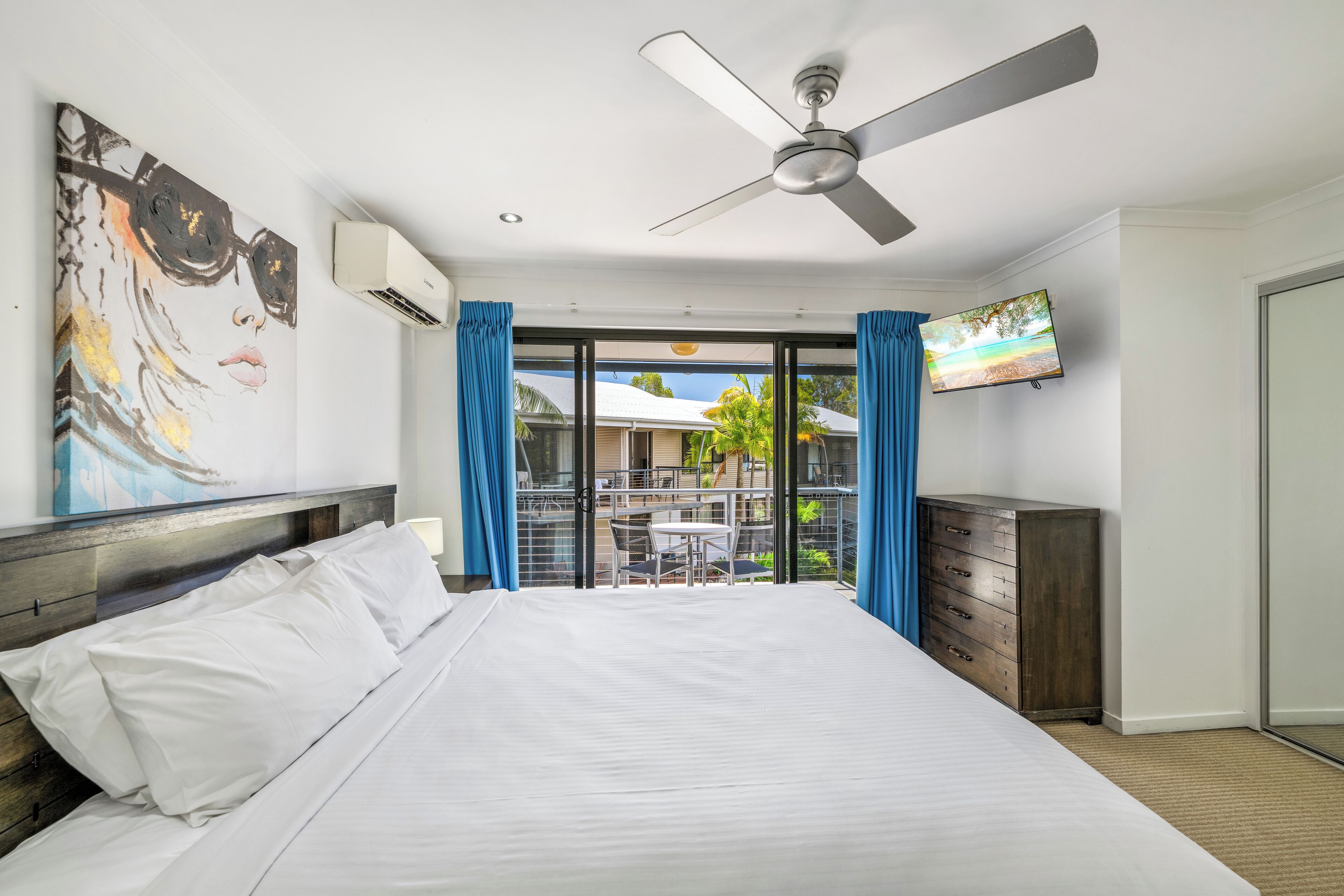 A hotel room with a large bed, a ceiling fan, a TV, and a balcony with a table and chairs.