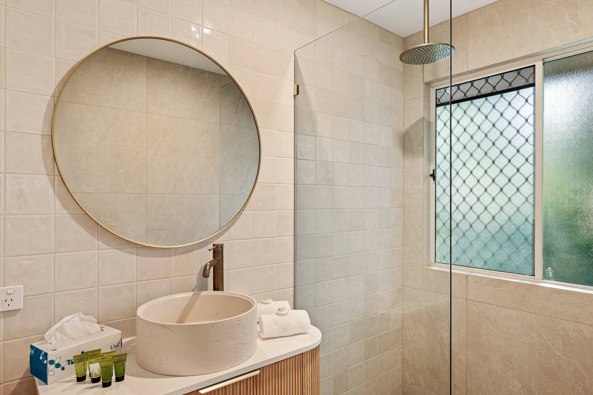 A bathroom with a round mirror, a sink, a showerhead, and a window with a frosted glass design.