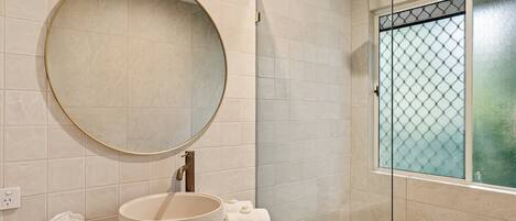 Un baño con un espejo redondo, un lavamanos, un cabezal de ducha y una ventana con un diseño de vidrio esmerilado.