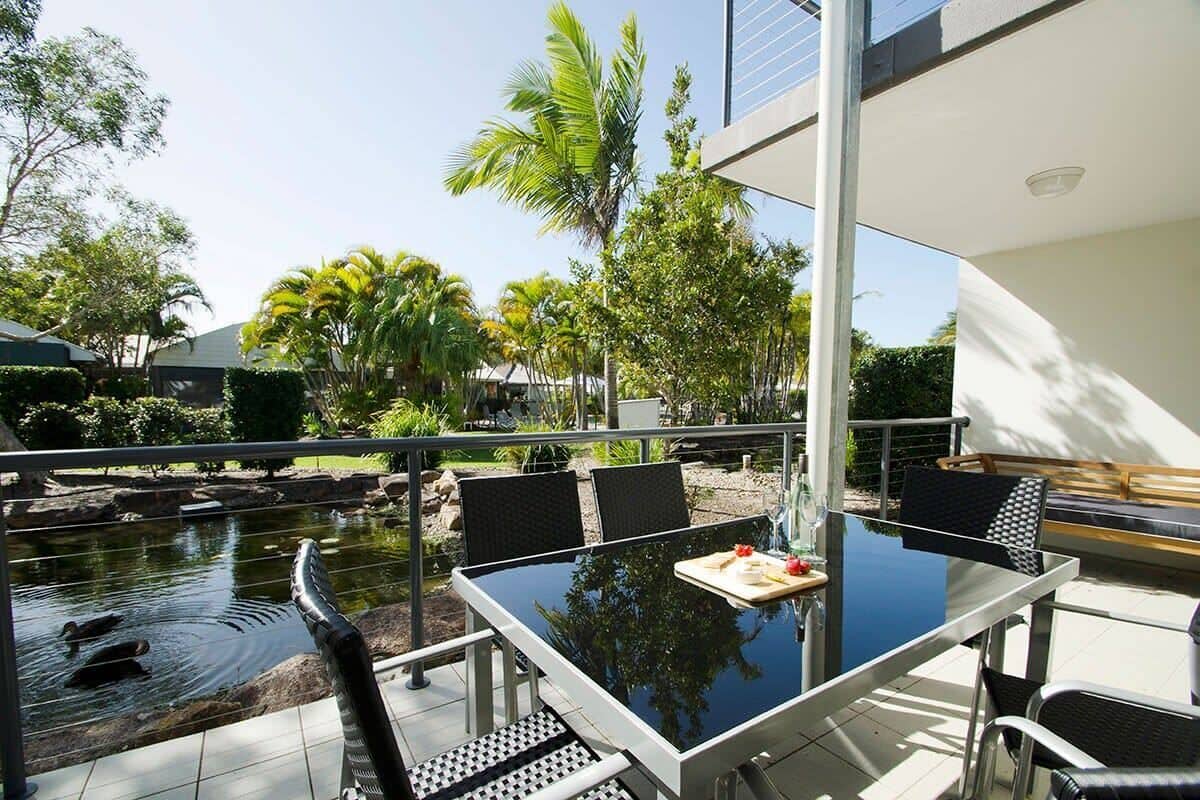 An outdoor dining area with a glass table and chairs, a small pond with ducks, and tropical plants.