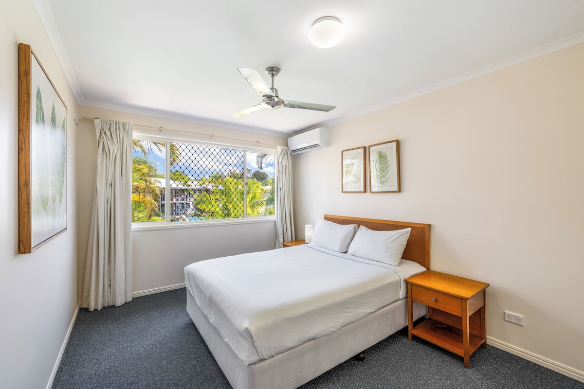 A hotel room with a bed, a nightstand, a ceiling fan, an air conditioning unit, and a window with curtains.
