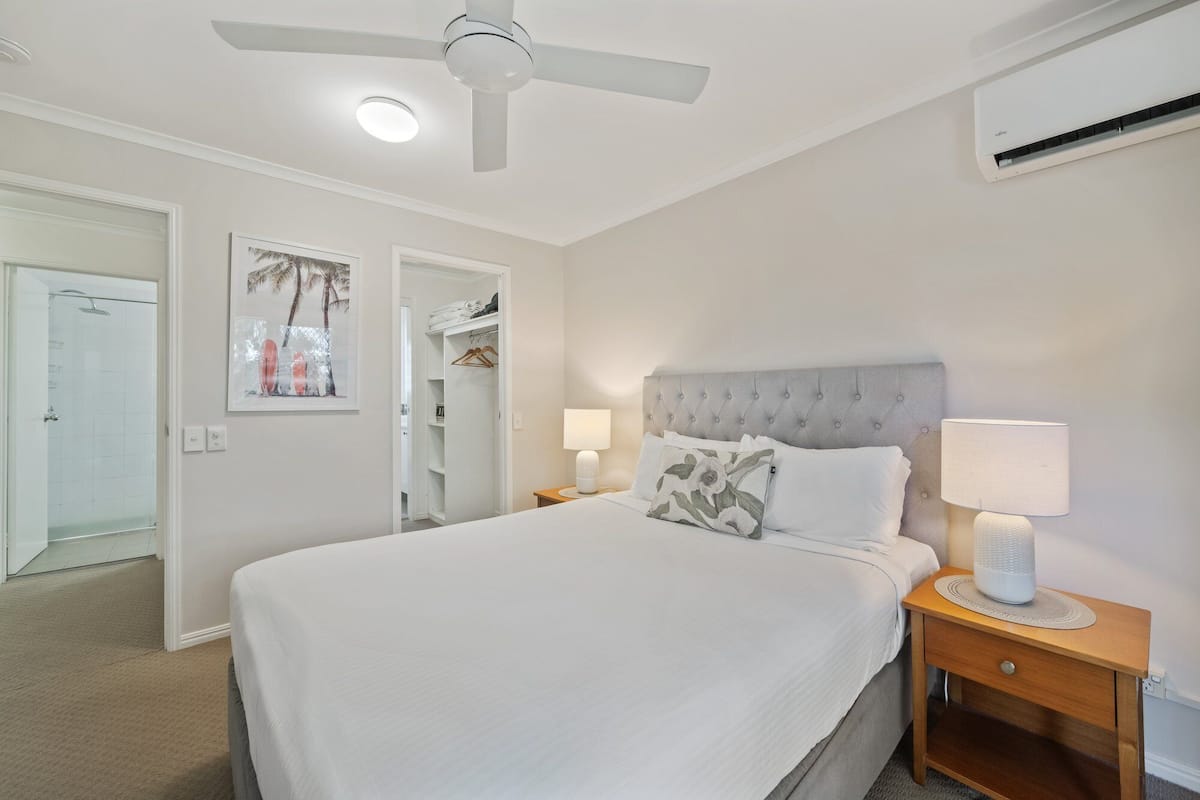 A bedroom with a bed, bedside lamps, a ceiling fan, an air conditioning unit, and a closet.
