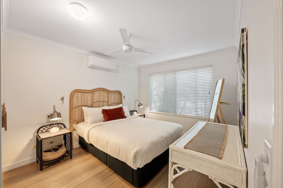 A bedroom with a bed, bedside table, lamp, ceiling fan, window with blinds, and an easel.