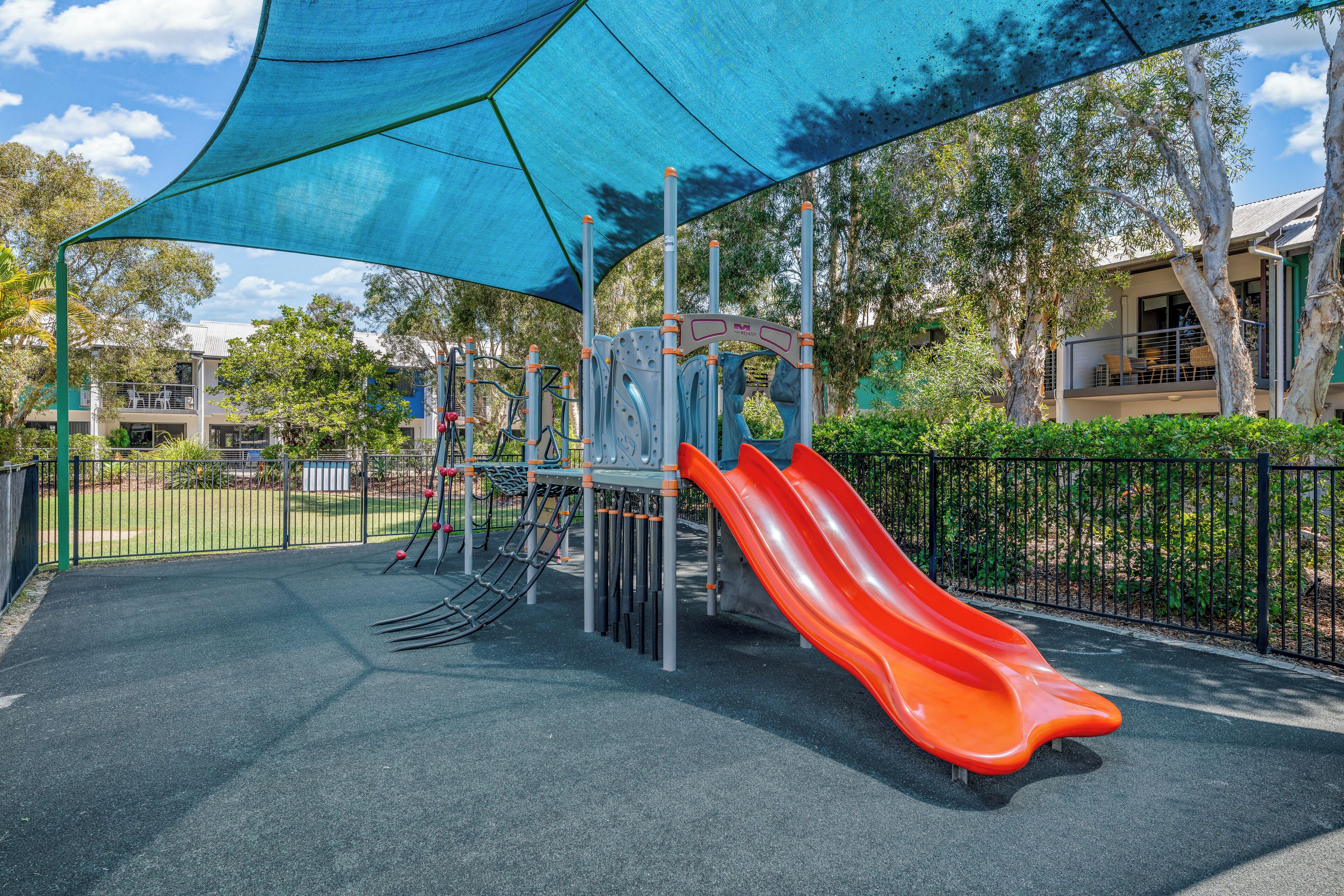 A playground with a slide and climbing structures, surrounded by a fence and trees.