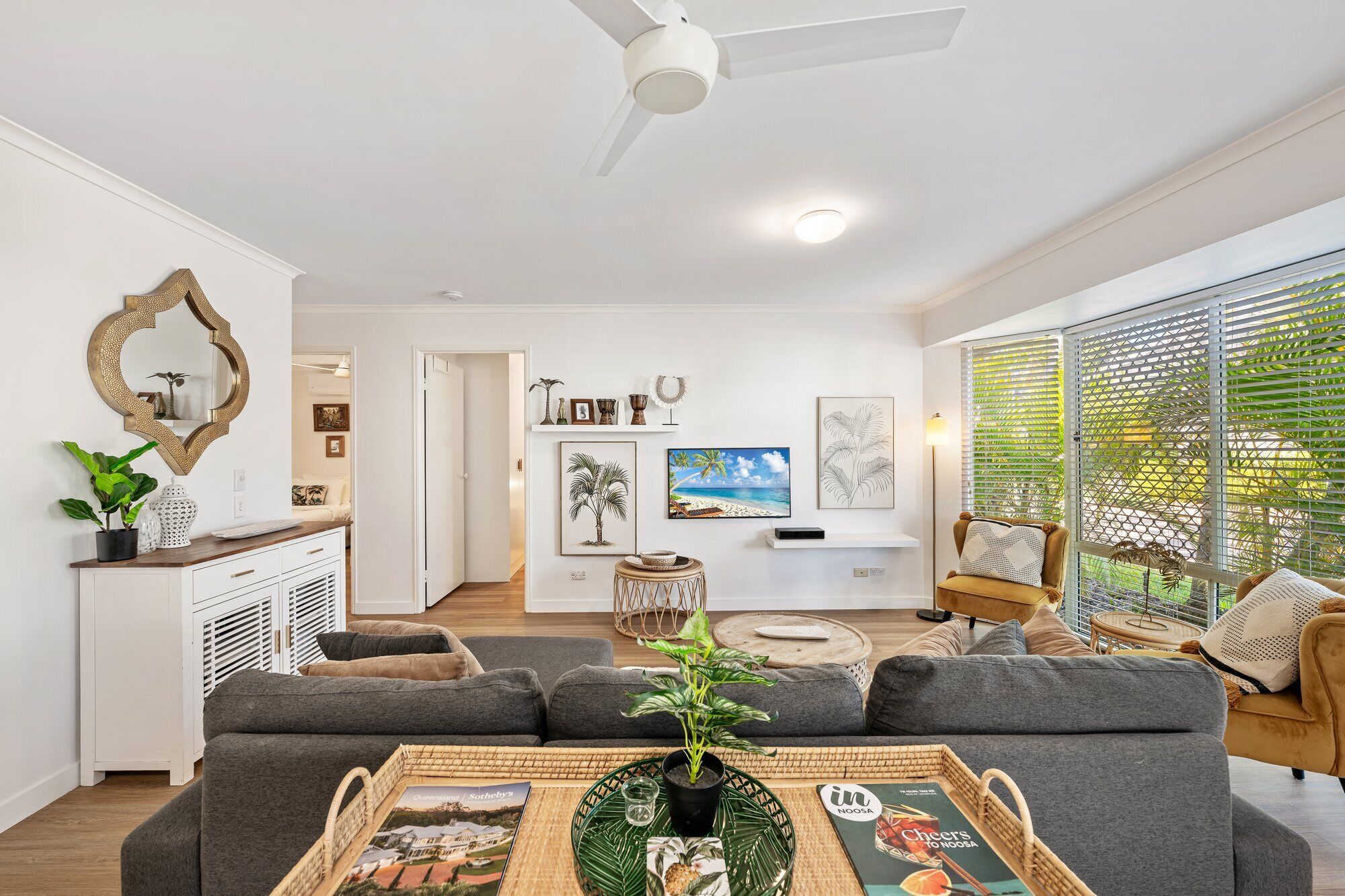 A modern living room with a sofa, a coffee table with magazines, a TV, and a dining area with a white cabinet and decorative items.