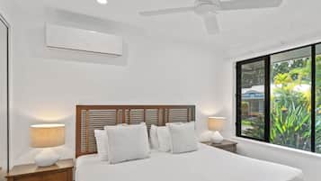 A bedroom with a large bed, wooden headboard, two bedside tables with lamps, an air conditioning unit, and a window with a view of greenery.