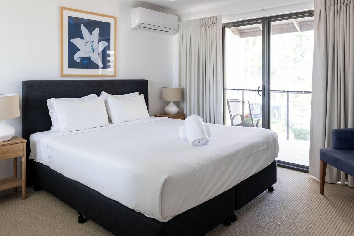 A neatly made bed with white linens and a rolled towel, a bedside table with a lamp, a framed picture on the wall, and a balcony with curtains.