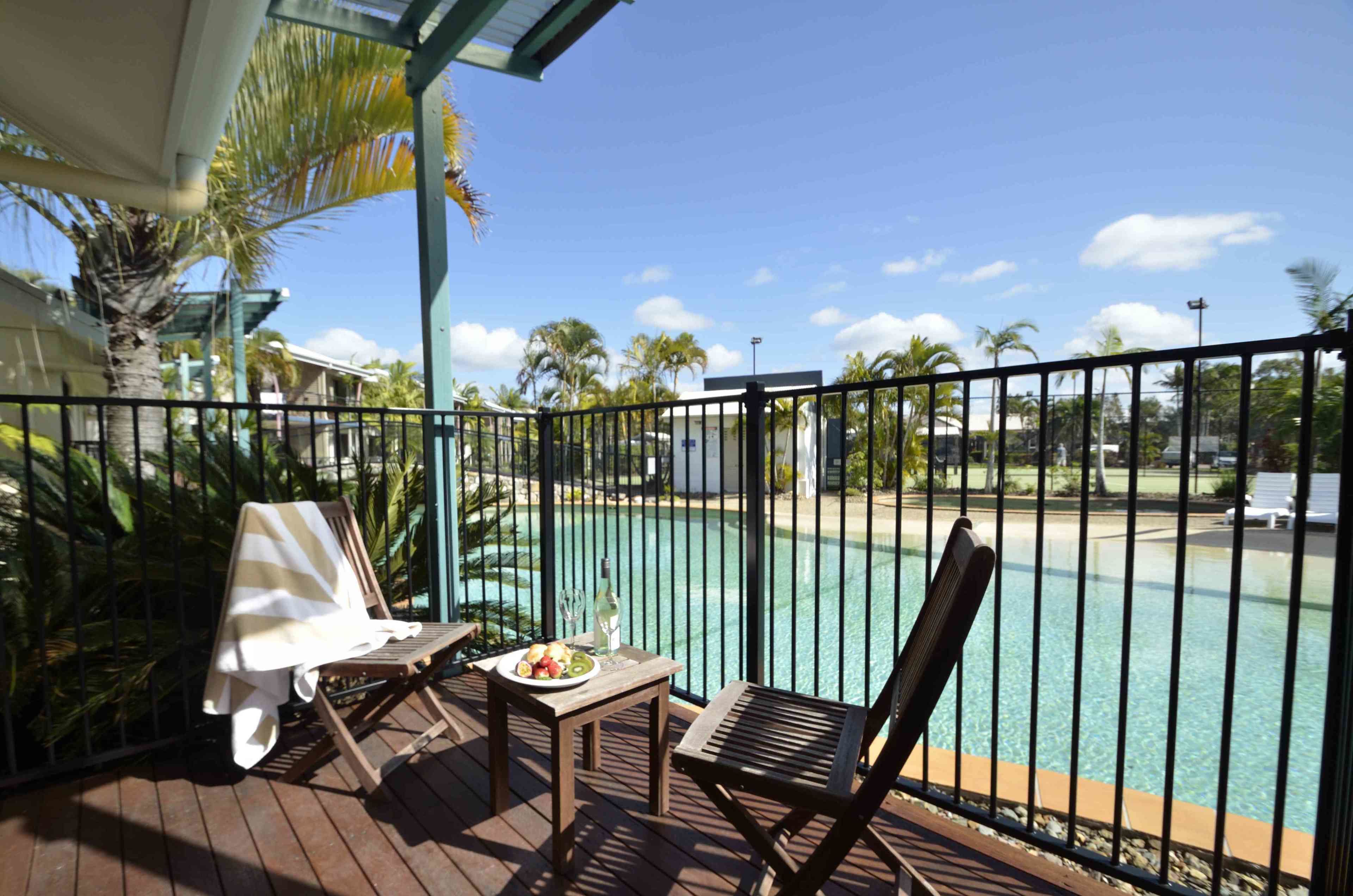 Un balcón con vista a la piscina, mobiliario de madera y una mesa con una fuente de fruta y una botella.