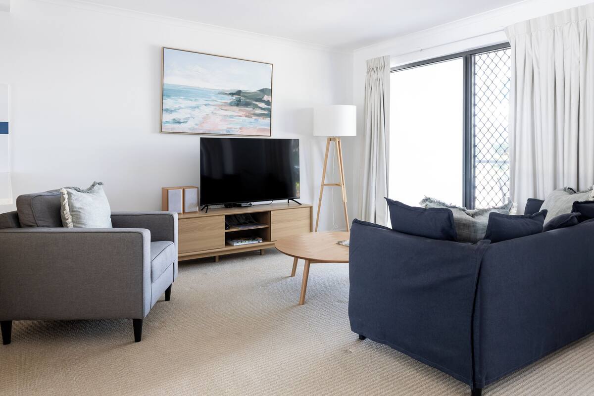 A modern living room with a sofa, a TV on a wooden stand, and a large window with curtains.