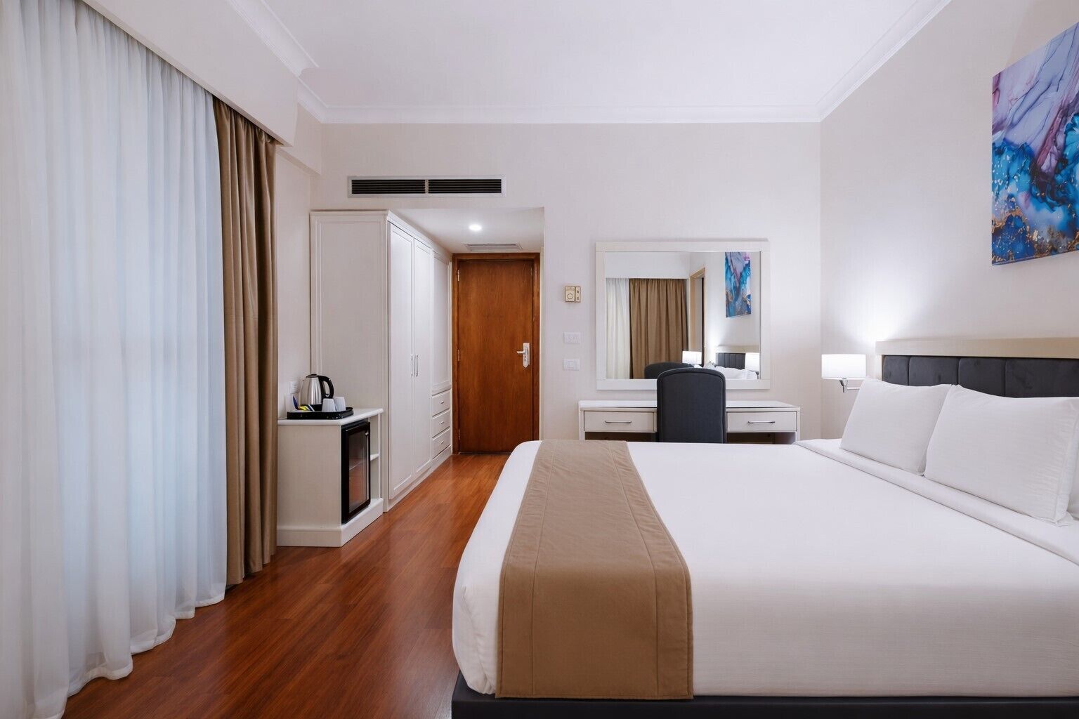A hotel room with a bed, a desk, a chair, a mirror, and a painting on the wall.