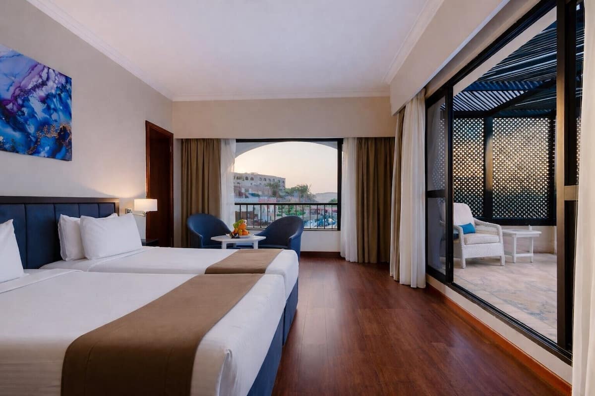 Habitación de hotel con dos camas, un balcón con vistas y una zona de comedor con sillas.
