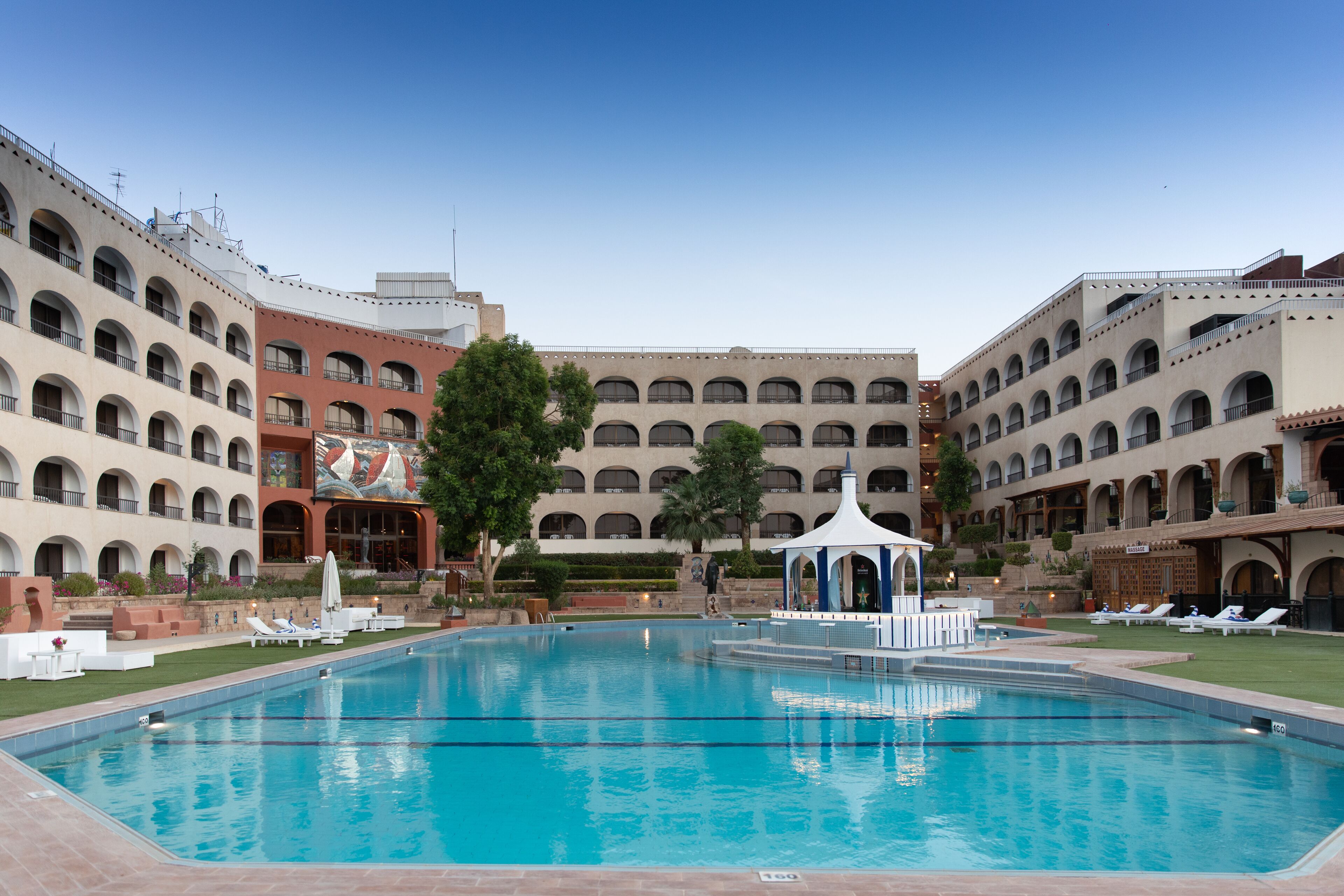 Una gran piscina con sillones de descanso y un pabellón, rodeada de edificios de varios pisos con ventanas arqueadas y balcones.