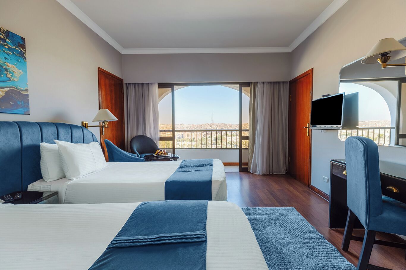 A hotel room with two beds, a desk, a chair, a TV, and a balcony with a view.