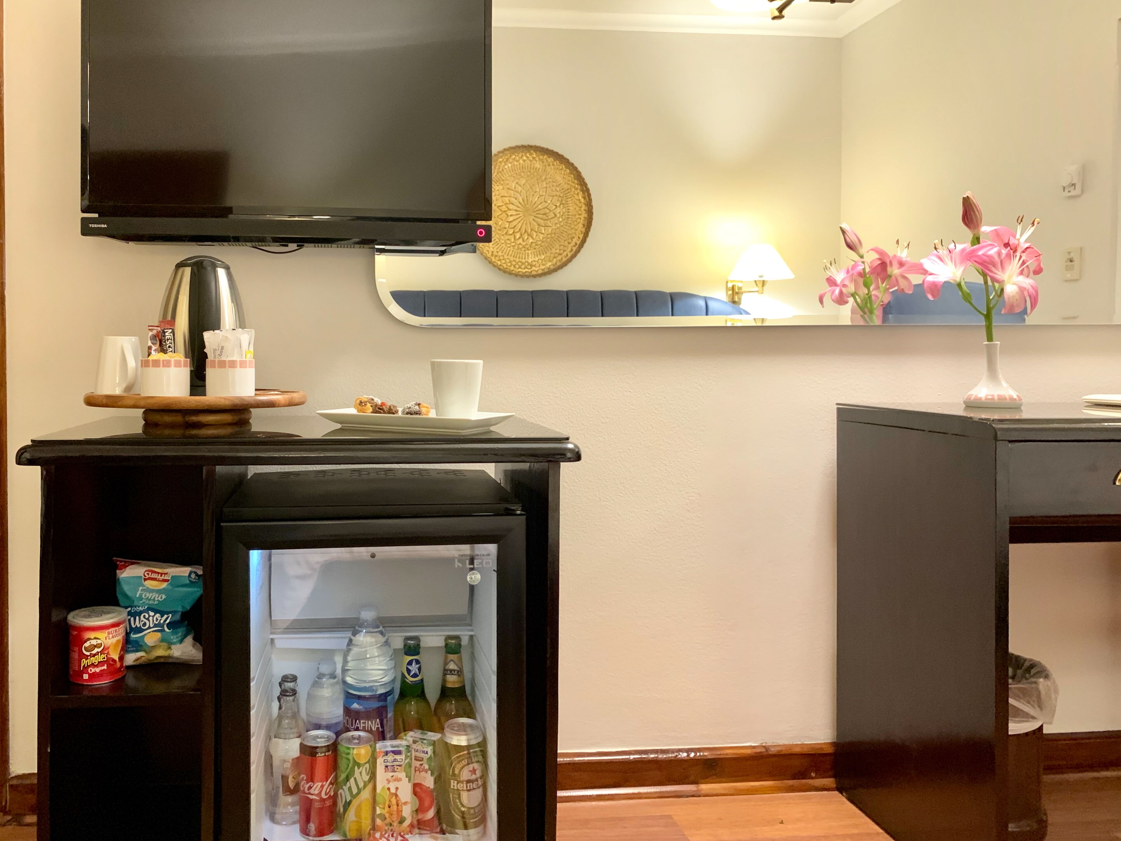 Habitació d'hotel amb televisió de pantalla plana, minibar amb aperitius i begudes, taula petita amb làmpada i un ram de flors.