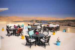 An outdoor seating area with round tables and chairs, potted plants, and a view of a desert landscape with mountains and a body of water.