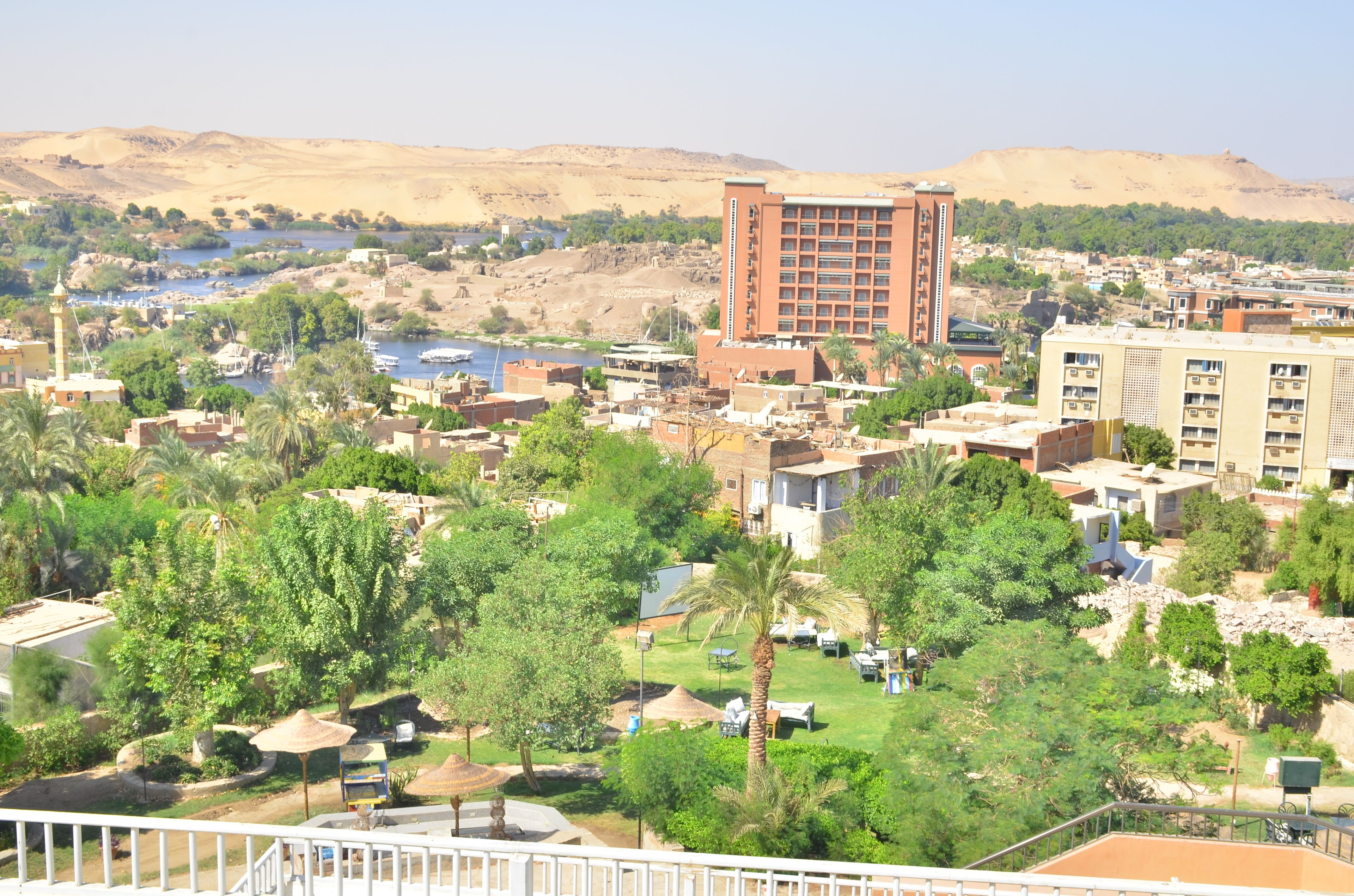 A hotel with multiple floors, a central courtyard with palm trees, and a view of the surrounding landscape including hills and a body of water.