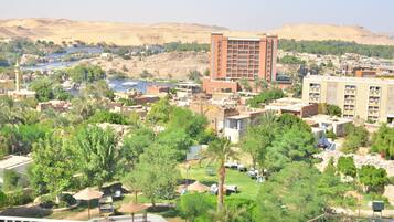 A hotel with multiple floors, a central courtyard with palm trees, and a view of the surrounding landscape including hills and a body of water.