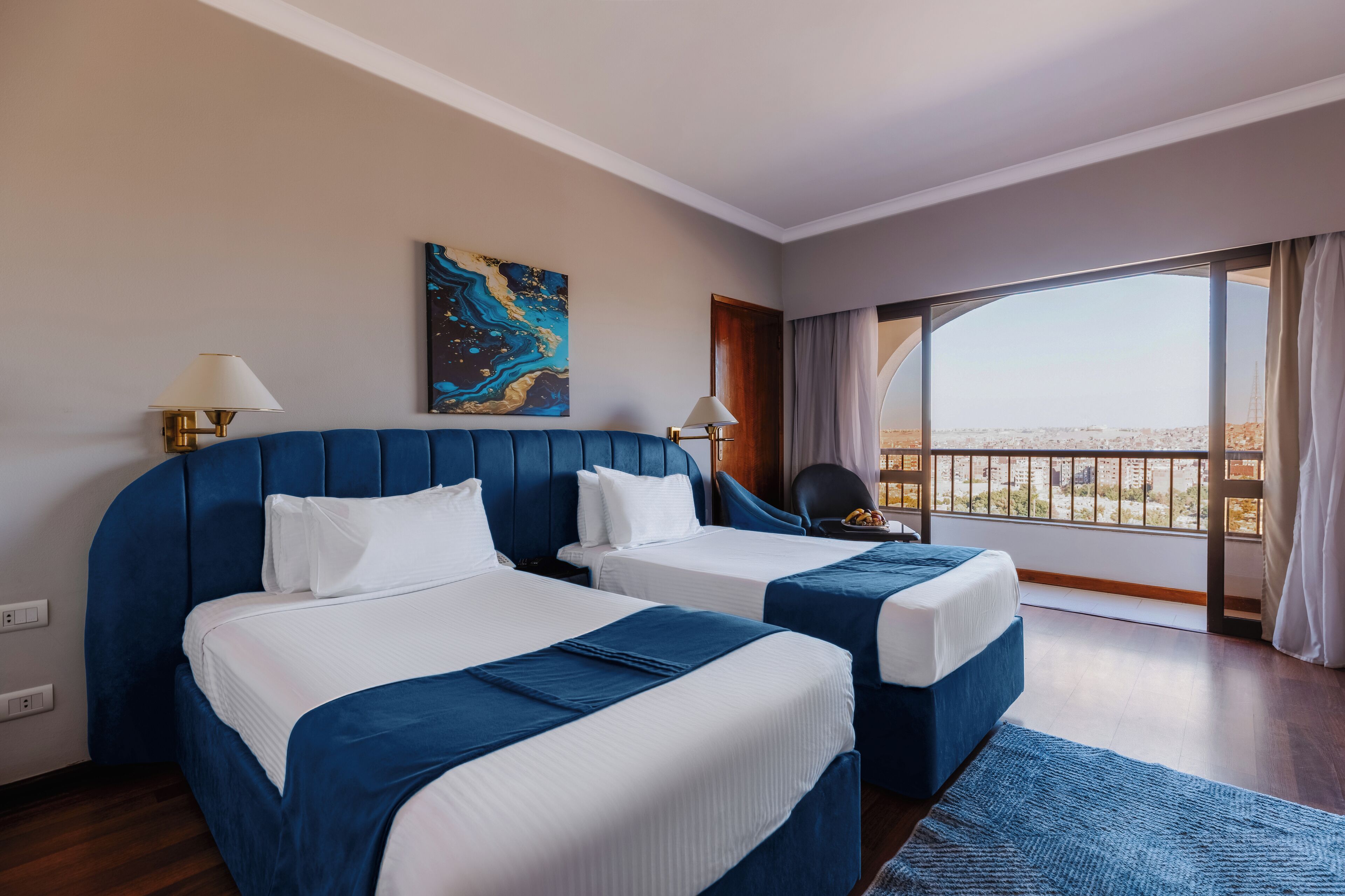 A hotel room with two beds, a large window with a view, and a painting on the wall.