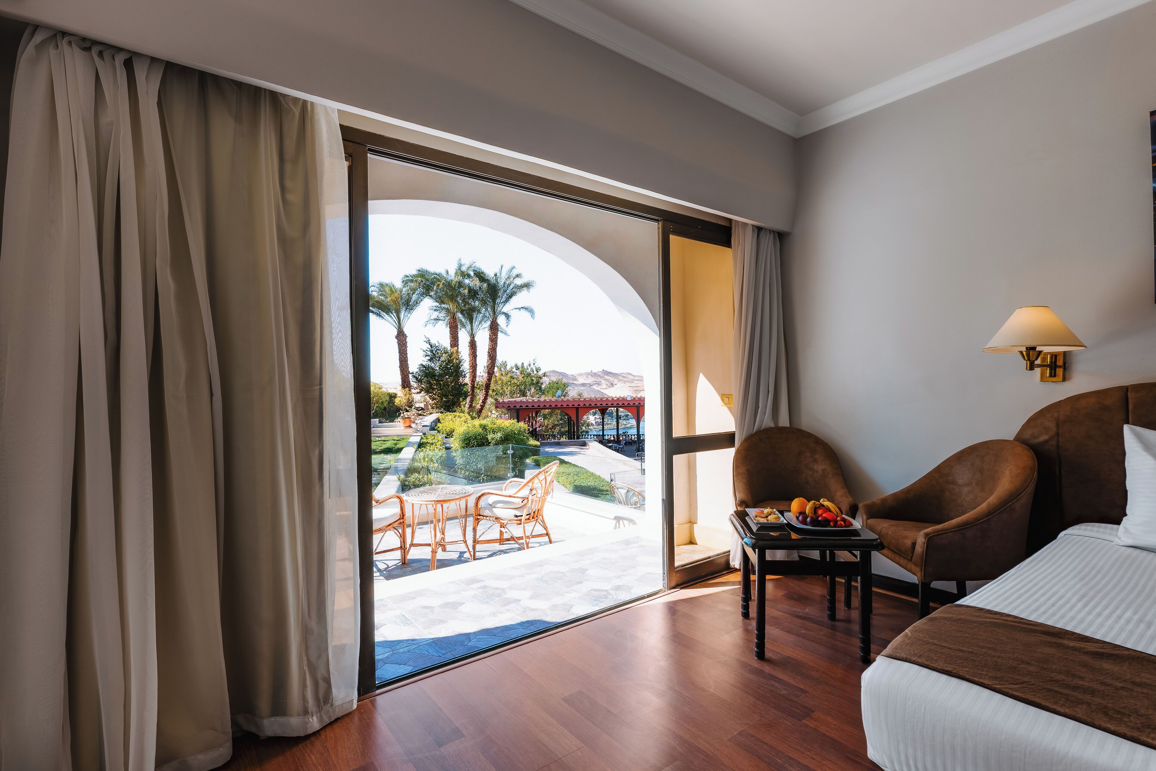 Habitación de hotel con un gran ventanal que da a un espacio exterior con comedor y palmeras.